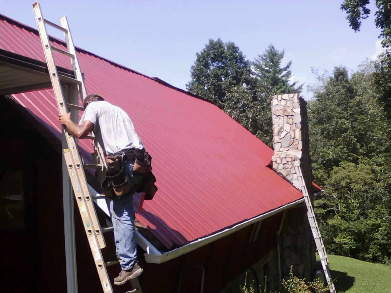 Roofer on a ladder beside a red metal roof and stone chimney — Soffit & Fascia Repair by Roofing Brothers in North Hollywood